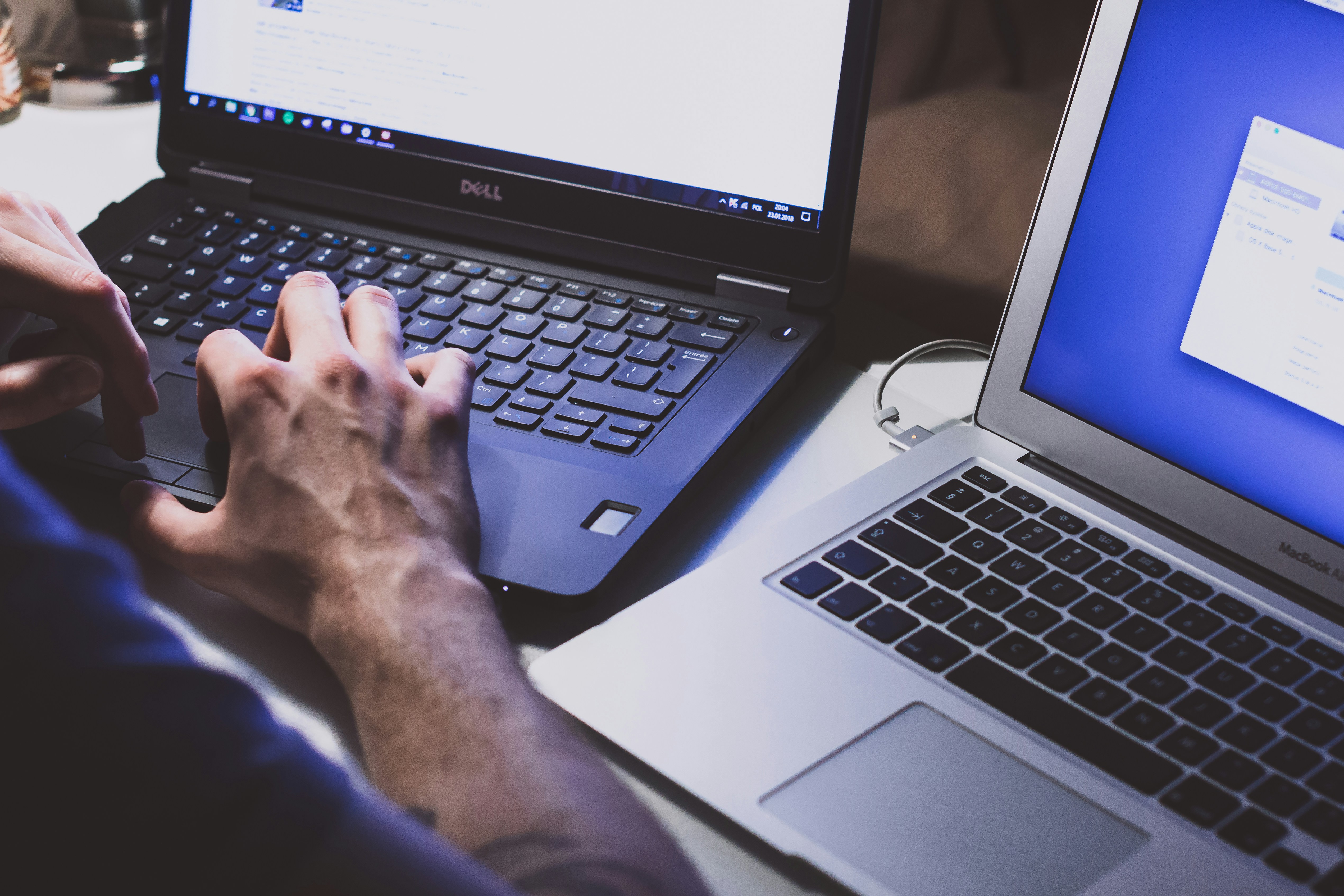 A person typing on a Dell laptop beside a MacBook, with dim lighting highlighting the hands and keyboards.