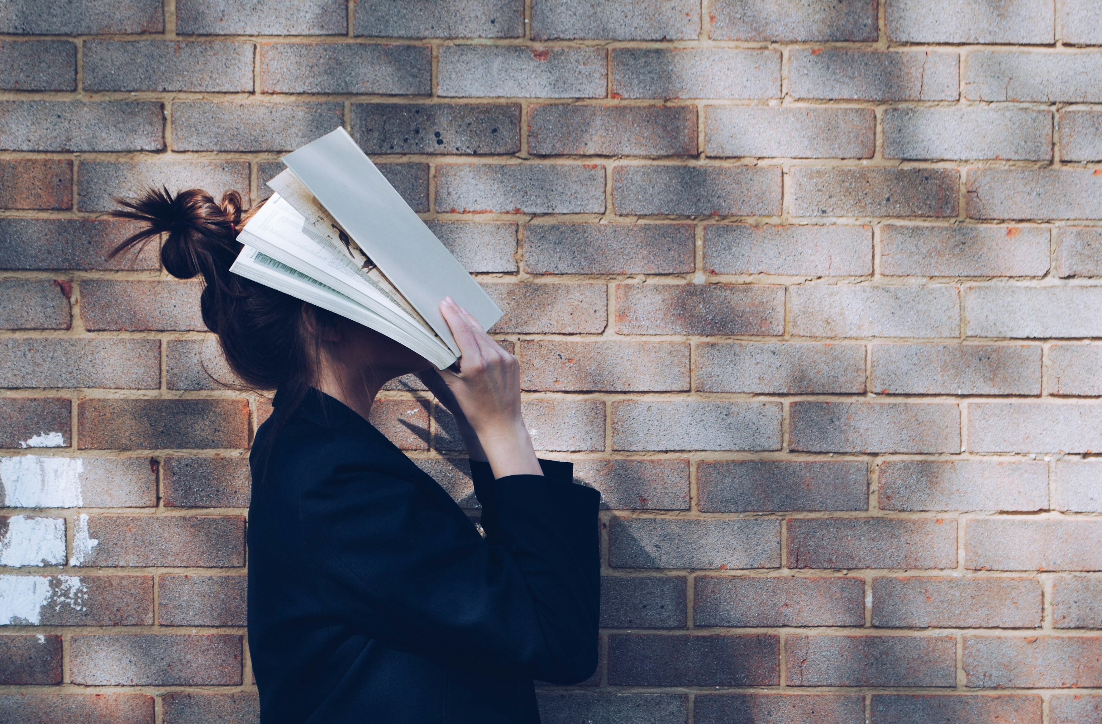 A student in a dark jacket holds an open book over their face against a brick wall, conveying stress or frustration.