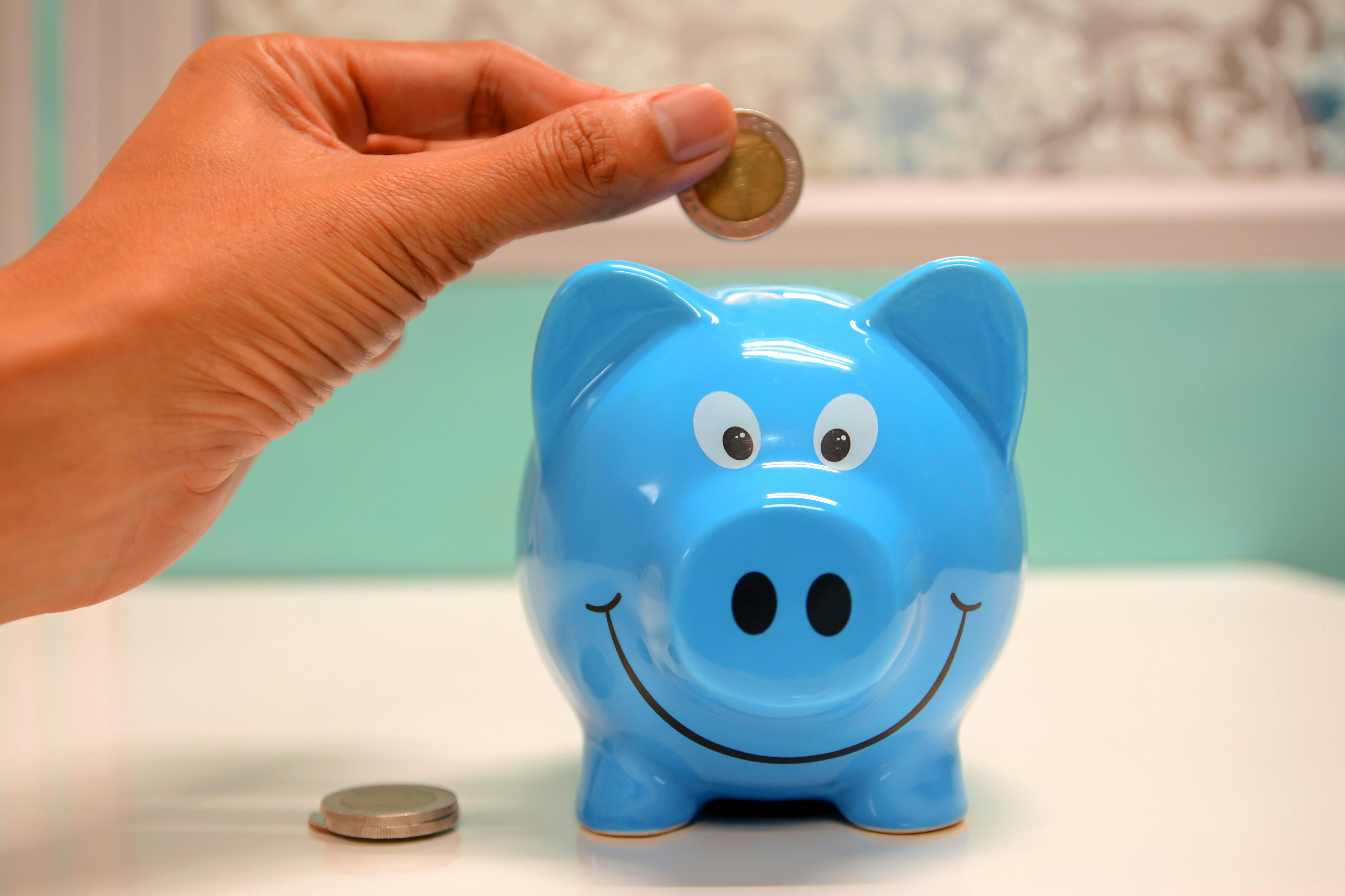 A close-up of a hand inserting a coin into a blue piggy bank with a smiling face. A few coins are placed on a white surface beside the piggy bank.